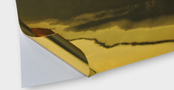 T74-P-P bright two-way gold metallised sign film showing reflections of light in the shiney bright surface. One corner of the film is folded back to reveal the release liner which clearly shows the two-way viewing nature of this film, as the adhesive side looks exactly the same metal gold colour as the glossy front side.