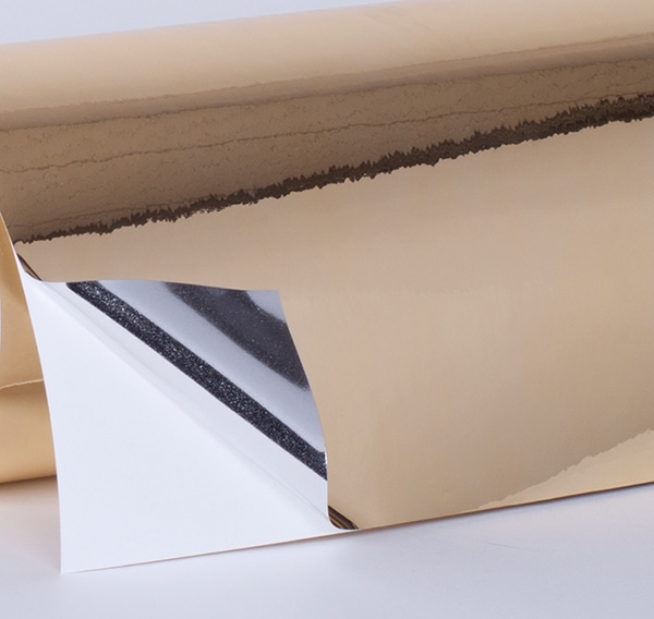 TSGG3 gloss gold one-way viewable metallised print film on a roll with one corner edge folded over to show the shiney silver colour back on the adhesive side of the film as it is pulled away from the liner.