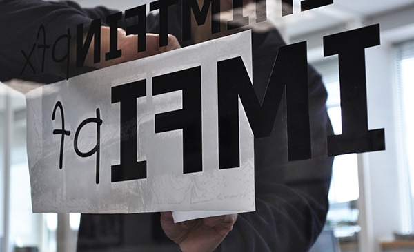 Transfering black cut vinyl lettering onto an internal window viewed through the glass you can see the application tape being peeled back and exposing the letters, as viewed from the adhesive side of the transfer paper.