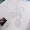 Applying TT60 vinyl graphics application tape over self-adhesive cut-vinyl graphics of a bear flying through the air, surounded by pollen seeds while holding an umbrella and a suitcase.