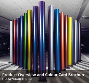 DOWNLOAD PDF VINYL COLOUR BROCHURE HERE: Avery Dennison Supreme Wrapping vinyl range of coloured rolls stood on-end in a garage with cars just visible in the background. The spectrum of coloured vinyl rolls are posed in an arrow shape pointing towards the centre of the photo.