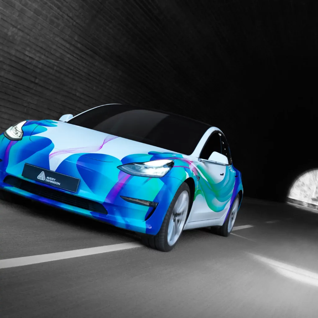 Avery Dennison SP 1504 EA non-PVC, conformable print wrap film is shown applied fully covering this car in a blue, pink and white swirling, water-like graphic design and the vehicle is speeding through a dark tunnel with daylight in the background.
