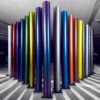 The photo shows a selection of the Avery Dennison Supreme Wrapping Films range of self-adhesive colour change vinyls for vehicle wrapping projects, with 17 coloured rolls standing vertically in their protective roll end-caps in a garage with cars just visible in the background. The spectrum of coloured vinyl rolls are posed in an arrow shape pointing towards the centre of the photo.