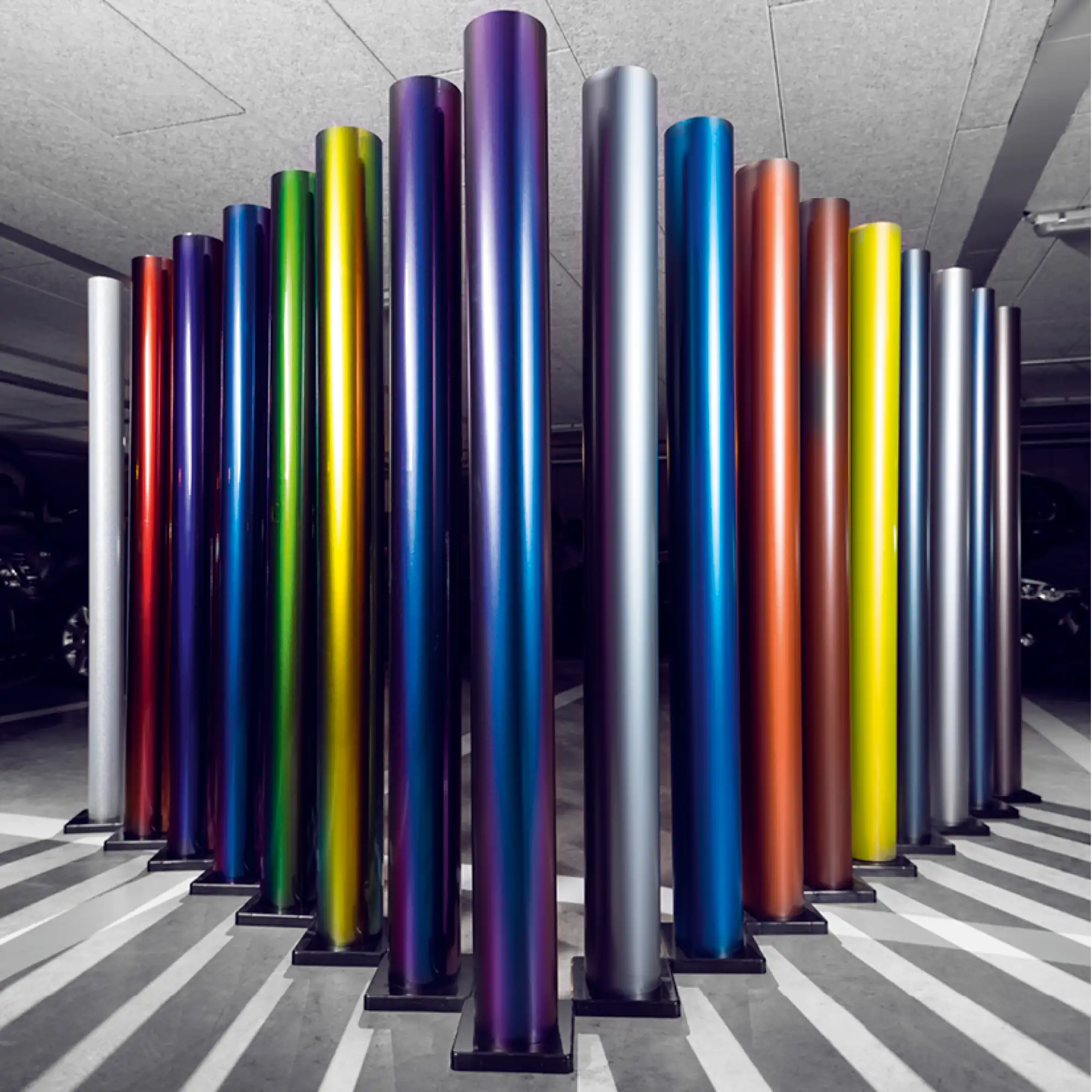 The photo shows a selection of the Avery Dennison Supreme Wrapping Films range of self-adhesive colour change vinyls for vehicle wrapping projects, with 17 coloured rolls standing vertically in their protective roll end-caps in a garage with cars just visible in the background. The spectrum of coloured vinyl rolls are posed in an arrow shape pointing towards the centre of the photo.