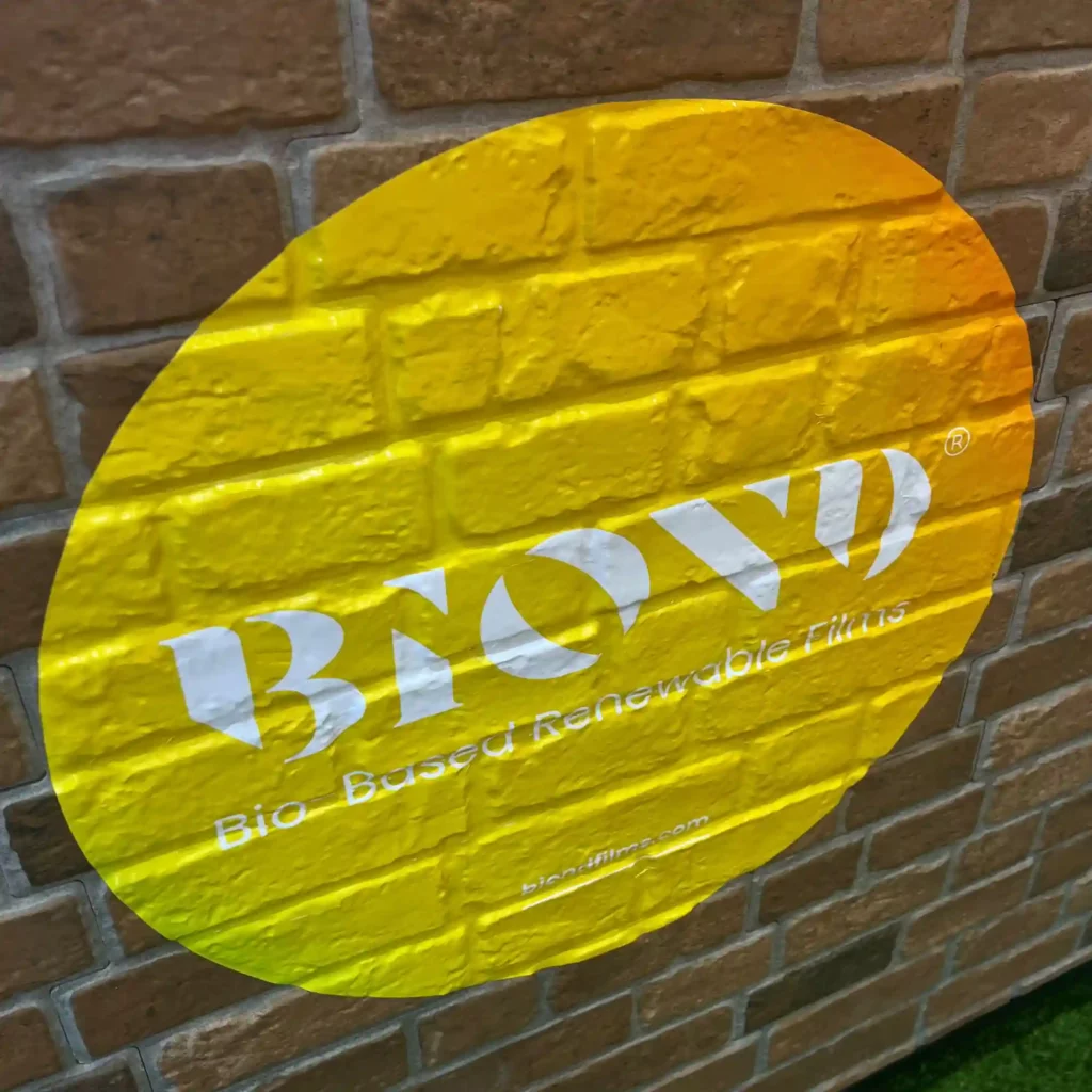 BIOND eco-friendly print film printed with a huge yellow cutout shape and renewable sustainable films text, applied onto an exterior textured brick wall.