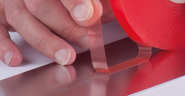 The outside (top) protective liner of DS-C-2065F clear double-sided sign tape is being peeled away to expose the adhesive on the top side as the tape on the other side (bottom) is adhered in a straight line to a Di-Bond metal sheet on a work bench.