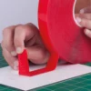 The outside (top) red protective liner of UHB-C-10F clear double-sided sign tape is being peeled away to expose the clear adhesive on the top side as the tape on the other side (bottom) which is being adhered in a straight line to a Di-Bond metal sheet on a graphics cutting mat.