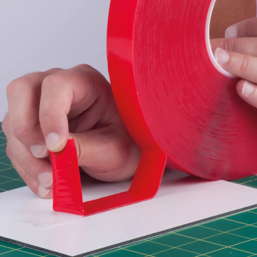 The outside (top) red protective liner of UHB-C-10F clear double-sided sign tape is being peeled away to expose the clear adhesive on the top side as the tape on the other side (bottom) which is being adhered in a straight line to a Di-Bond metal sheet on a graphics cutting mat.