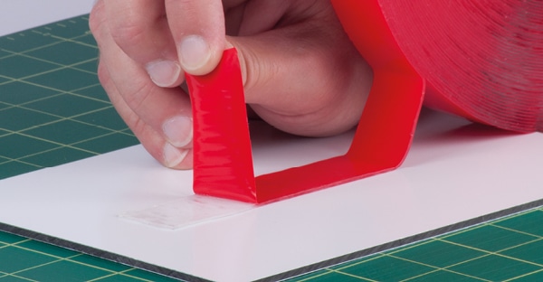 The outside (top) red protective liner of UHB-C-10F clear double-sided sign tape is being peeled away to expose the adhesive on the top side as the tape on the other side (bottom) is adhered in a straight line to a Di-Bond metal sheet on a work bench.
