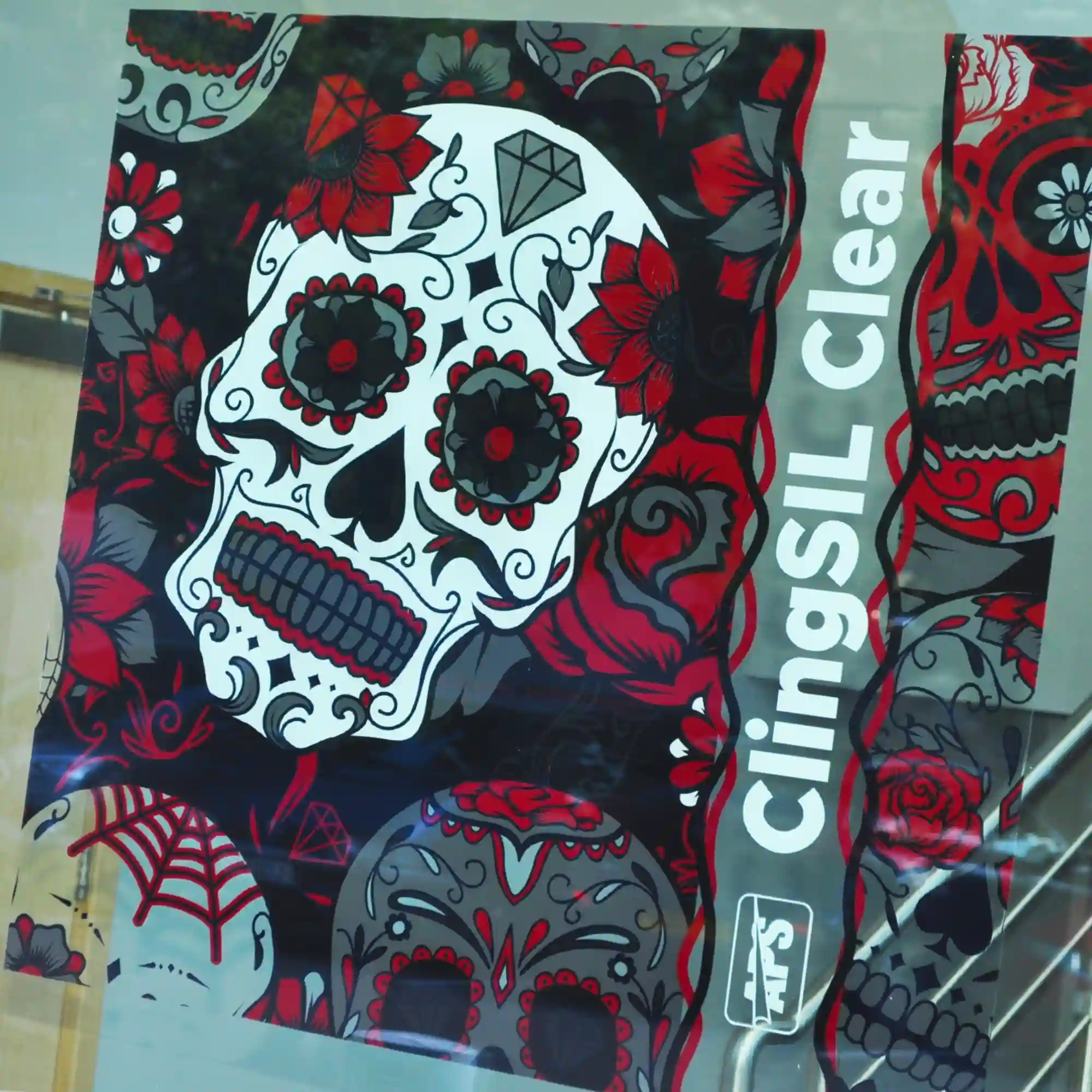 An ultra-clear gloss window cling film, ClingSil Clear CSC130, printed with a vibrant 'Day of the Dead' skull design. The colourful skull image is adorned with traditional Mexican decorative elements like floral patterns, intricate linework, and bright hues, with the transparent film allowing light to pass through while creating a festive and eye-catching window display.