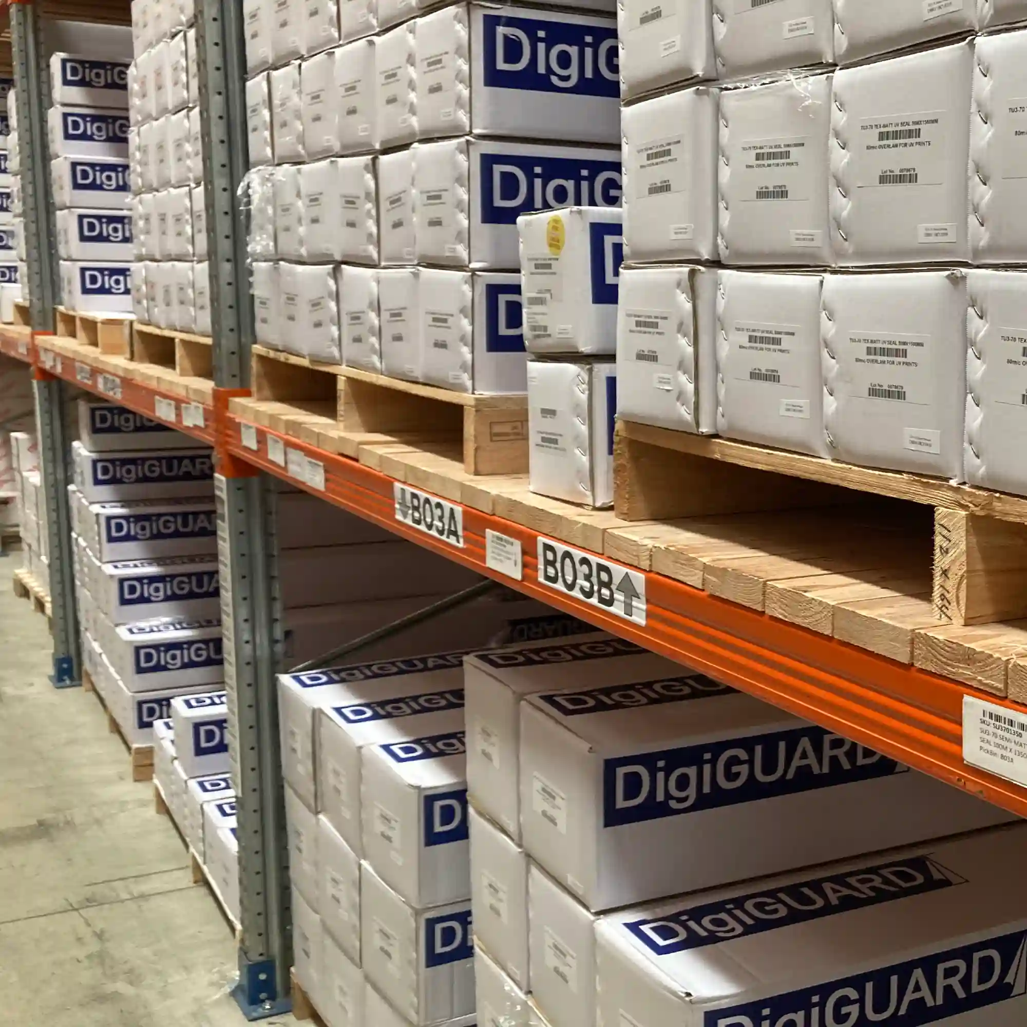 Textured matt graphics laminate, DigiGUARD TU3-70 for self-adhesive signage and UV digitally printed graphics, boxed in branded packaging across multiple levels of shelving within a warehouse aisle environment.