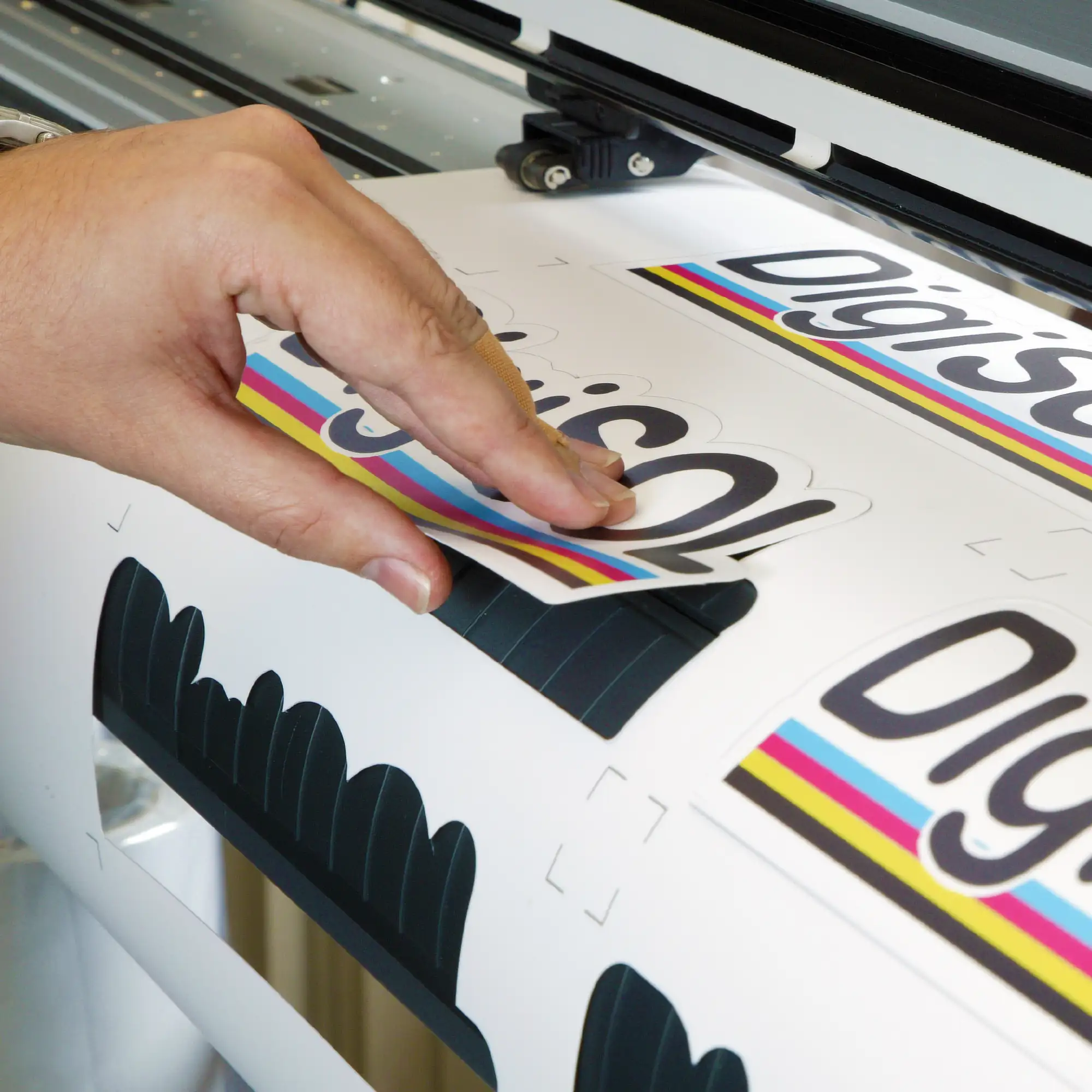 DigiPRINT DS3005, a thick and easy to apply print vinyl hangs with it's leading edge from the front of a wide format digital printer after the film has been printed with a brand logo and then plotter cut to produce fully cutout print n cut labels. One completed label is being removed from the imposed roll while it is hanging from the printer.