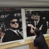 DigiPRINT DS3045 blockout gloss white PVC print film is in the process of being applied to an office glass window with the printed self-adhesive vinyl featuring a bold, high-contrast graphic of the iconic Blues Brothers in black suits, hats, and sunglasses standing in front of the soul food cafe with the abstract styled image looking sharp and clear and the film fully obscures the sun and the outside view from showing through the glass, providing complete privacy from the outside while displaying a classic, eye-catching, retro-inspired design on the inside of the glass. The glossy finish enhances the choice of dark colors and gives the print a polished, professional look.