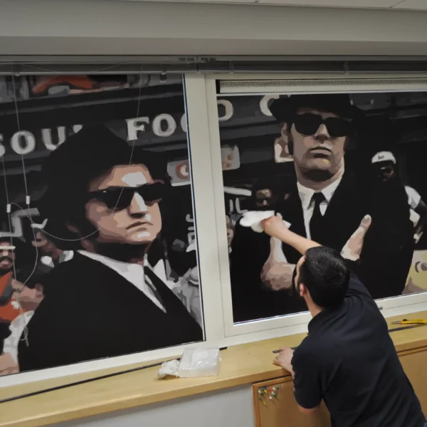 DigiPRINT DS3045 blockout gloss white PVC print film is in the process of being applied to an office glass window with the printed self-adhesive vinyl featuring a bold, high-contrast graphic of the iconic Blues Brothers in black suits, hats, and sunglasses standing in front of the soul food cafe with the abstract styled image looking sharp and clear and the film fully obscures the sun and the outside view from showing through the glass, providing complete privacy from the outside while displaying a classic, eye-catching, retro-inspired design on the inside of the glass. The glossy finish enhances the choice of dark colors and gives the print a polished, professional look.