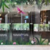 A store window features a vivid, full-color print using DigiPrint DS3054P permanent ultra-clear window film with graphics of bright tropical birds and vibrant jungle colors that stand out sharply against the transparent background, creating a high-impact visual without obstructing natural light. The ultra-clear film allows for a clean, frameless look, making the print appear as if it’s directly embedded into the glass. Designed for display prints onto glass surfaces, the permanent adhesive ensures the graphic remains securely in place, perfect for retail branding and decorative installations on windows.