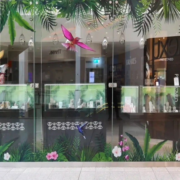 A store window features a vivid, full-color print using DigiPrint DS3054P permanent ultra-clear window film with graphics of bright tropical birds and vibrant jungle colors that stand out sharply against the transparent background, creating a high-impact visual without obstructing natural light. The ultra-clear film allows for a clean, frameless look, making the print appear as if it’s directly embedded into the glass. Designed for display prints onto glass surfaces, the permanent adhesive ensures the graphic remains securely in place, perfect for retail branding and decorative installations on windows.