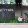 A full-color print using DigiPrint DS3054P permanent ultra-clear window film applied onto a glass store-front window with graphics of bright tropical birds and vibrant jungle foliage that creates a high-impact visual without obstructing natural light of the view through the glass into the store behind. The ultra-clear PVC film offers a good alternaive to clear polyester films when applying onto glass and allows for a clean, seamless 'no vinyl' look, making the print appear as if it’s directly embedded into the glass. Designed for display prints onto glass surfaces, the permanent self-adhesive secures the graphic in place and the clarity of the vinyl is perfect for retail branding and decorative installations on windows.