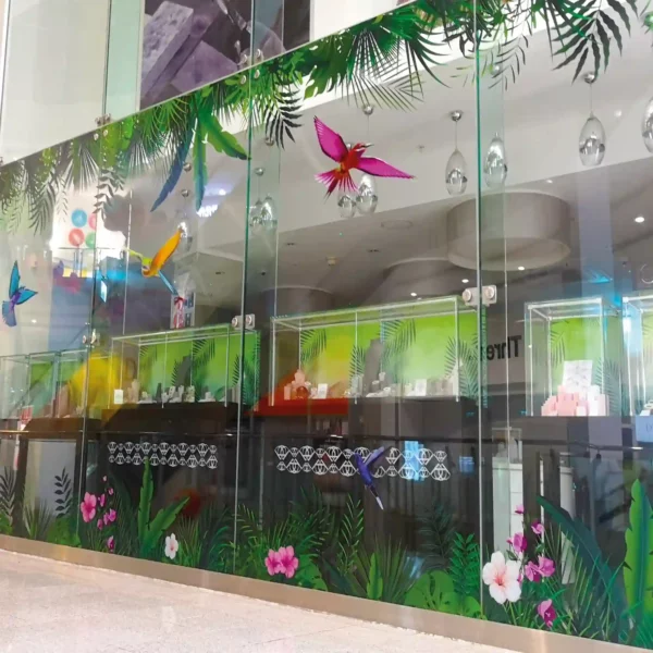 A retail storefront window features a vibrant, full-color graphic of tropical birds perched among lush green foliage, printed on ultra-clear vinyl. The image appears almost as if it's floating on the glass, thanks to the DigiPrint DS3054R, known as the clearest print vinyl for windows. The vivid hues of the jungle scene stand out brilliantly, while the transparent background allows natural light to filter through. This seamless, high-impact design draws attention without obstructing visibility, making it ideal for retail display windows.