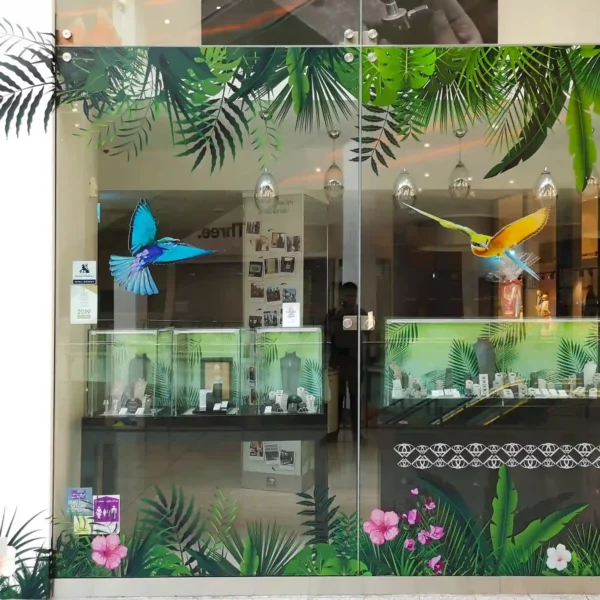 retail store window showcases a face-on view of a striking printed graphic featuring bright tropical birds, applied using DigiPRINT DS3054 R removable ultra-clear window print vinyl. The self-adhesive vinyl clings seamlessly to the glass, creating a vivid, high-resolution image that appears to float on the surface. The ultra-clear background allows full visibility through the unprinted areas, making the birds and foliage pop in brilliant color without blocking light or views. Perfect for promotional or seasonal displays, the vinyl is both removable and eye-catching, offering an elegant, see-thru branding solution for storefronts.