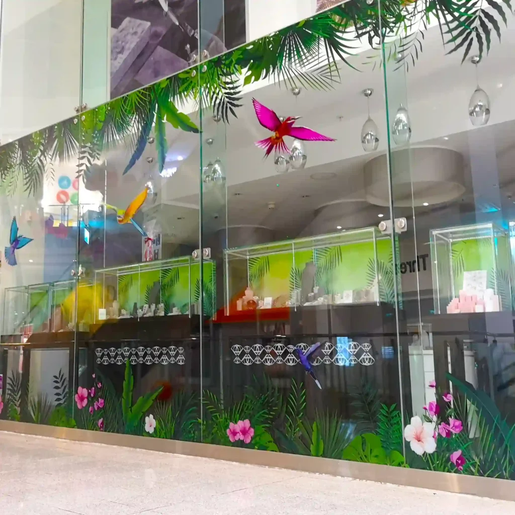 A retail store front with large glass windows is shown, where the glass surfaces are covered with vibrant, high-resolution printed jungle, parrots and colourful flower graphics applied to the glass, showcasing the "DigiPrint Speciality Window Films" series of self-adhesive printable vinyls. A bright and professional setting, that emphasizes the decorative functions of this series of window print films.