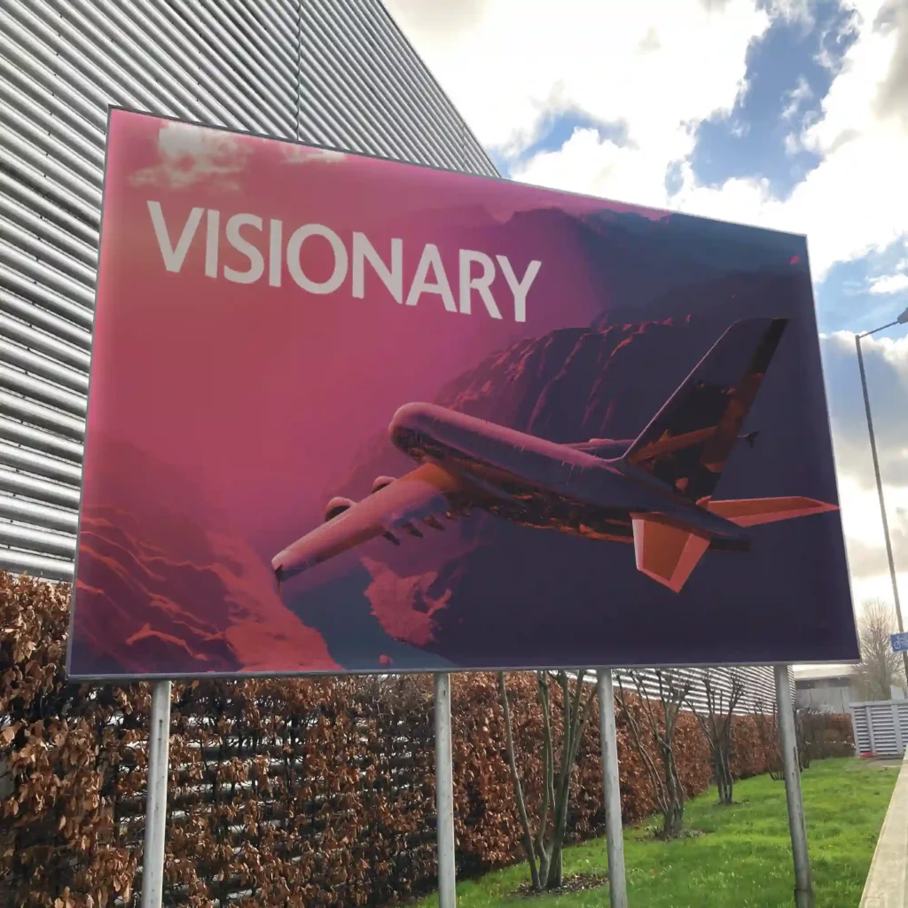 Impactful outdoor signage, using digitally printed billboard poster paper for a large format advertisement on a trading estate roadside that features 'Visionary' text and an image of a red sepia tinged aeroplane flying over mountains.