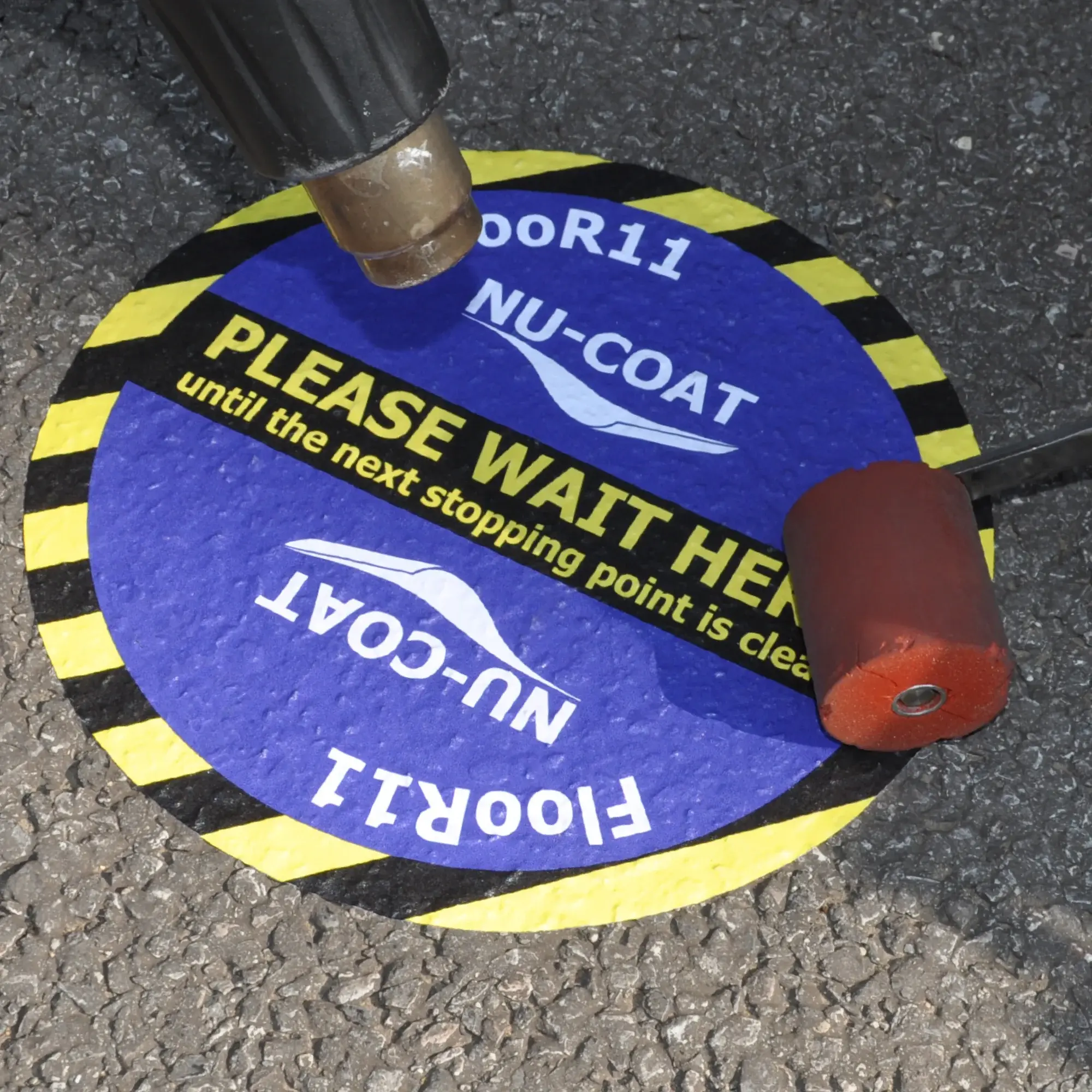 Slip rated floor print laminate, M77-P laminated to M15-M print vinyl and being applied with a heat gun and soft roller to an external tarmac surface for use as a directional wayfinding graphic with additional promotional floor advertising. Photo detail shows the vinyl confirmed into the rough gravel texture road surface.