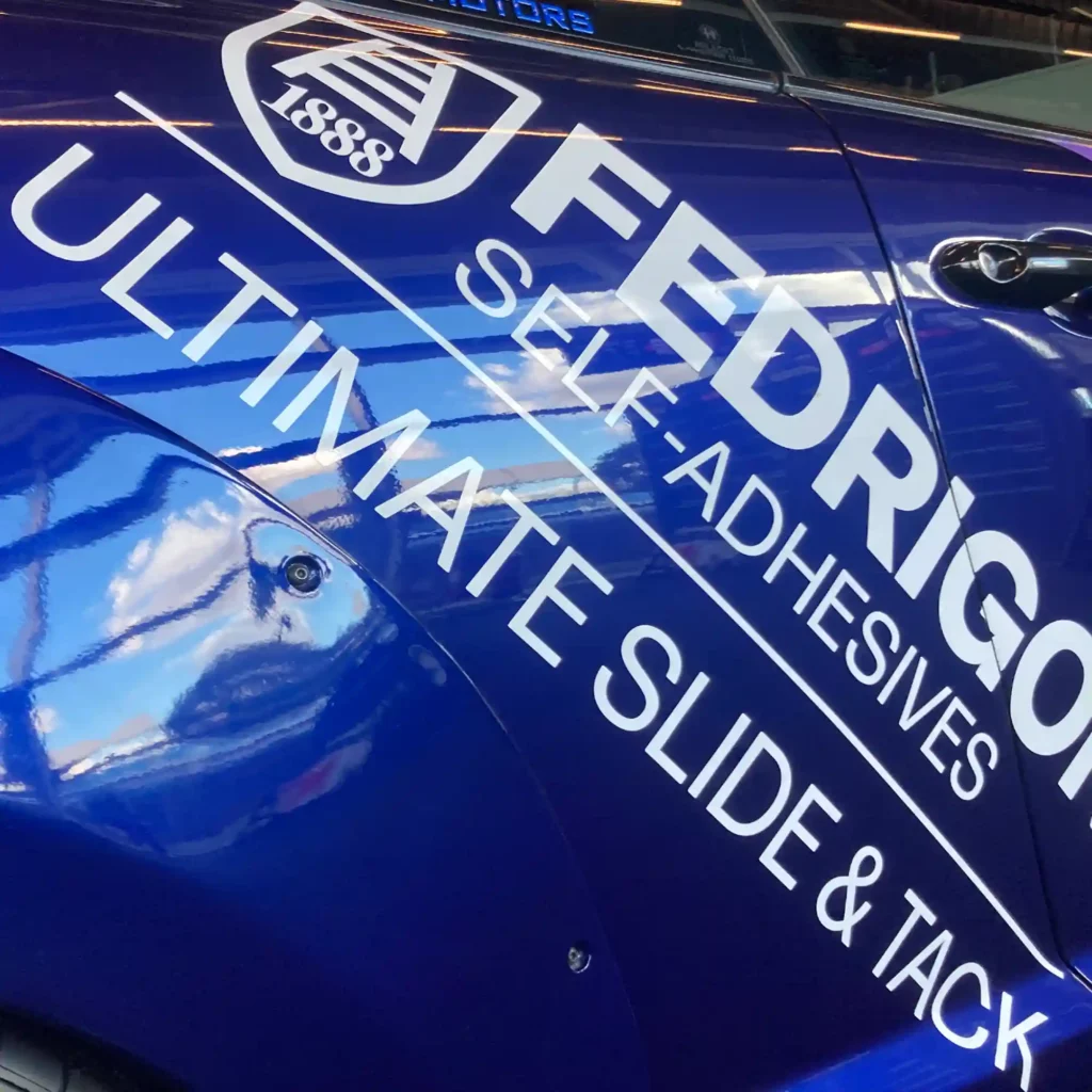 Fedrigoni Ultimate Wrappnig Film (UWF) gloss blue colour vehicle wrap film clode up on the side of a car with overlaid gloss white CAD-cut vinyl lettering applied over the top.