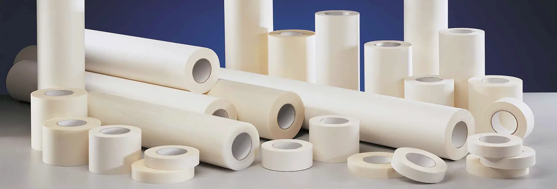 An assortment of graphics application tape papers for self-adhesive graphics transfer which have been slit to various roll log widths, all viewed side by side and stacked-up on a large table top.