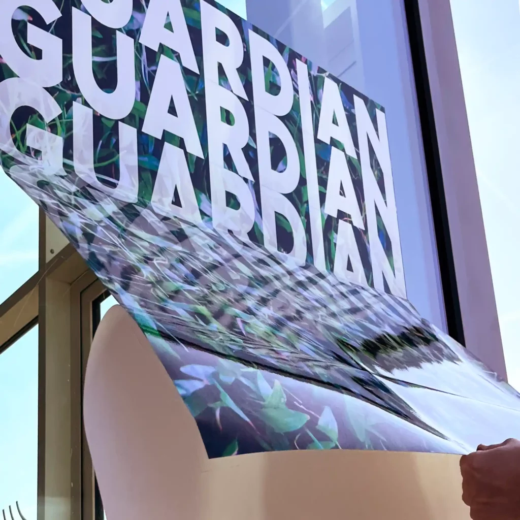 GUARDIAN Lam Gloss, non-PVC laminate for protective covering of digital prints, being applied to an exterior window as part of a short-term printed promotional signage project, shown with the self-adhesive prints' liner being pulled back, mid-installation, so it can be stuck onto the application surface.