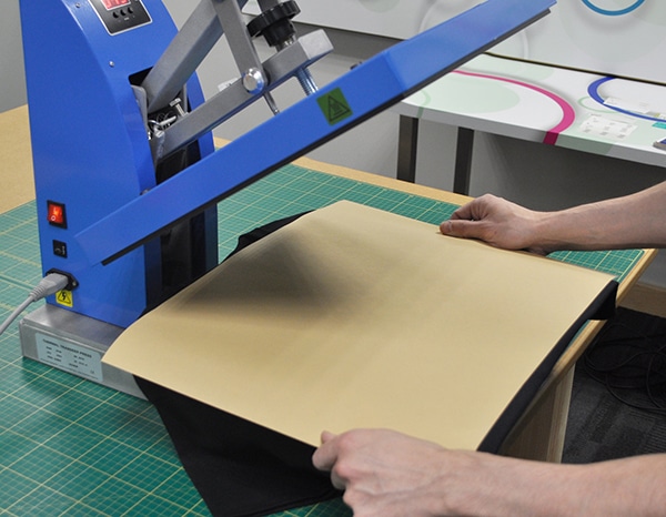 An open heat press (essentially a large flat iron on a hinge!) with a t-shirt under it between the two heated elements and a brown Cover Sheet is being placed on top of the clothing to protect the vinyl garment decoration film being applied and to protect the heat press.