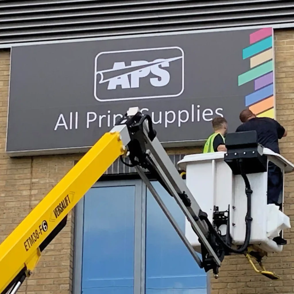 JT5826 P speciality self-adhesive printing film for applications onto plasticised banners being applied, with the aid of a cherry picker, over an existing plasticized sign material that is situated under the high eaves of the All Print Supplies office building.