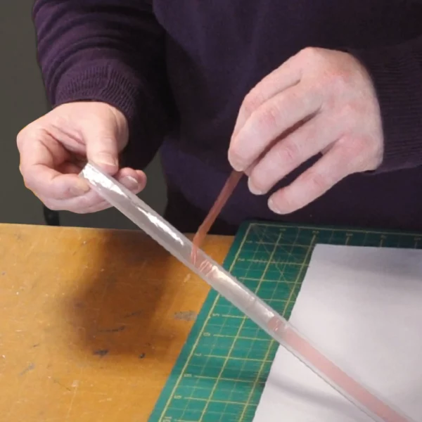 Peeling off the protective red adhesive liner to reveal the sticky, self-adhesive side of the Keder PVC self-adhesive framing tape used for inserting and holding printed textile graphics into tension display framing systems.