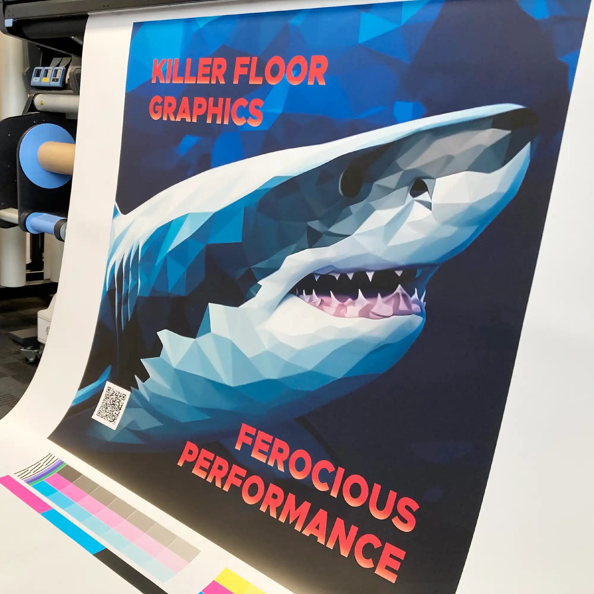 Kernow FloorSharK, self-adhesive R10 slip rated floor graphic material in the process of being printed on a wide format digital ink printer, with a large shark graphic protruding from the printer bed surrounded by red text stating "KILLER FLOOR GRAPHICS... FEROCIOUS PERFORMANCE".