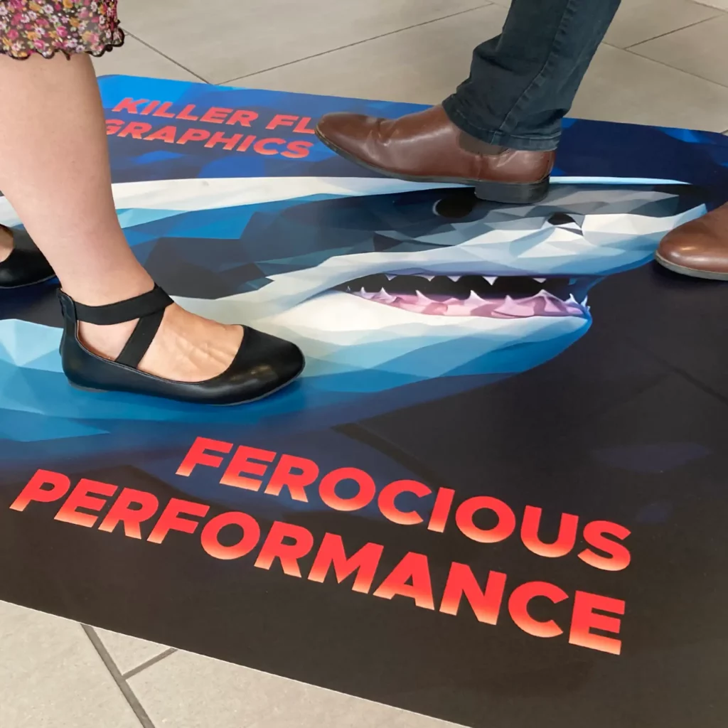 Kernow FloorSharK being walked over after being applied onto interior tiled floor surface. The printed graphic features a stylised image of a Great White Shark with red bold text surrounding the graphic, saying "Killer Floor Graphics - Ferocious Performance".