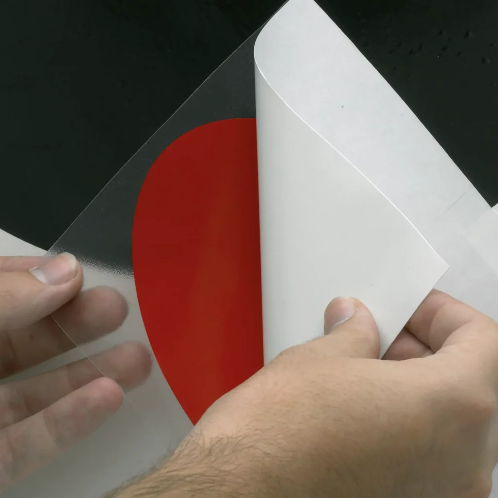 Clear application tape for self-adhesive graphics, KleenView KV40 is pictured with a red cutout circle shaped sign vinyl left attached to the transparent transfer film while the release liner is peeled away to expose the adhesive side of the vinyl prior to positioning and final application.