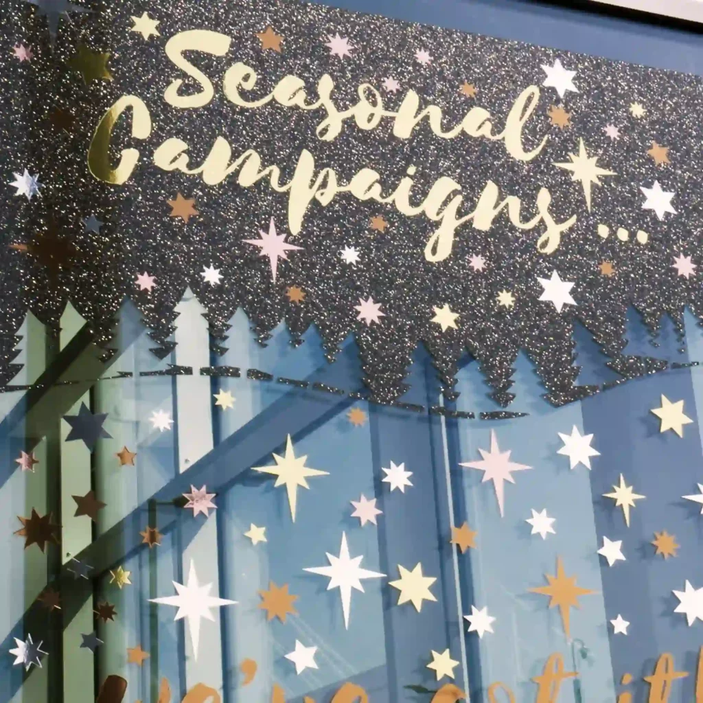 A large format seasonal campaign window graphic poster featuring vinyl colours from the KPMF K75100 Glitter Series of self-adhesive films, including the K75150 Black Glitter colour which is used to create the "starry night" background which the other golds, silvers and bronze metallic cut-vinyl text and sparkling shining stars are placed on top of to create this special promotional signage with a luxurious and premium look.