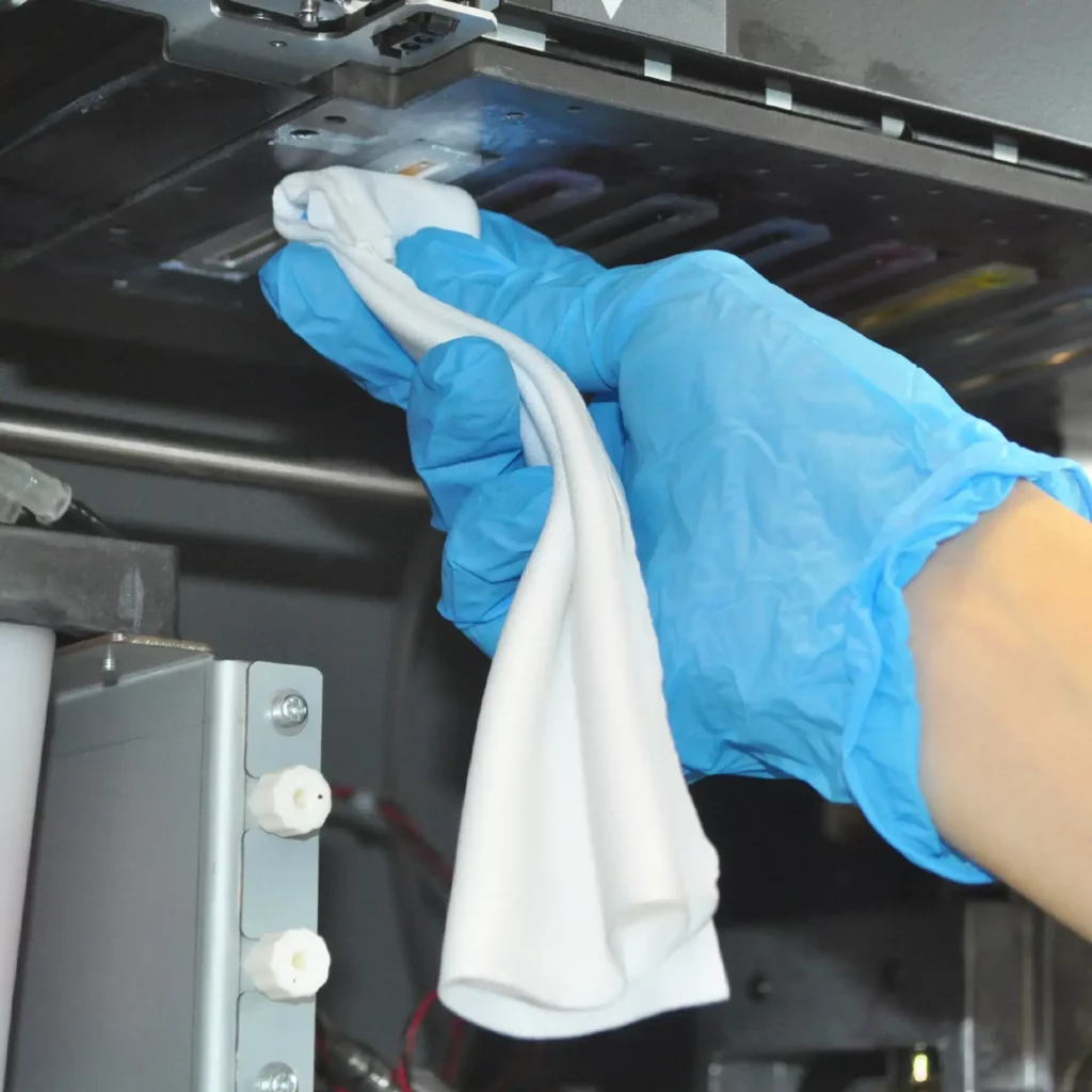 A white lint free cloth has been folded into the fingers and is being held in hand with a blue surgical glove while being used to clean around the print head of a wide format Solvent inkjet printer.