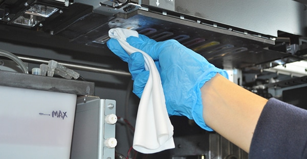 A white lint free cloth has been folded into the fingers and is being held in hand with a blue surgical glove while being used to clean around the print head of a wide format Solvent inkjet printer.