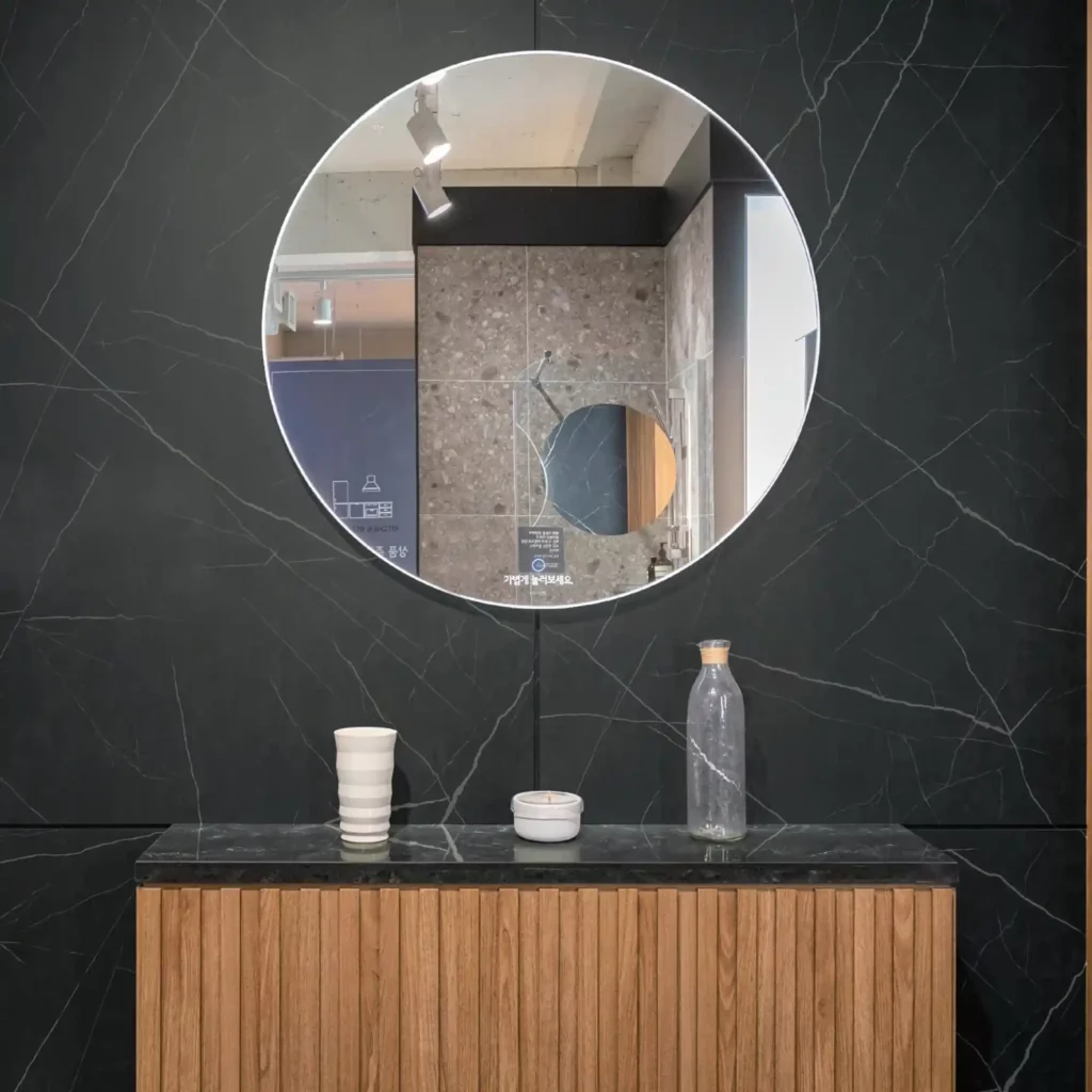 LX Hausys Interior Films 'BM' range of finishes decorate this elegant washroom wall and counter surface. The wall is entirely covered with a bold white veined black marble patterned self-adhesive vinyl matching the counter top and below that a complimentary slatted wood pattern effect film and a very large circular mirror is hung on the wall above the counter top.