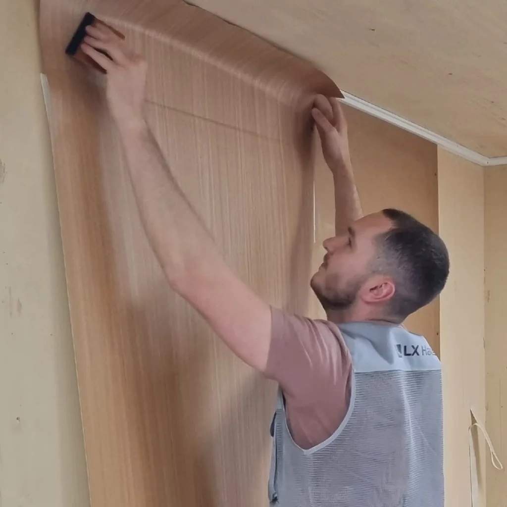 A light coloured wood effect LX interior film being applied with a squeegee onto a primed wall surface, working from the top of the wall before fully pulling the liner away as working way down the wall.