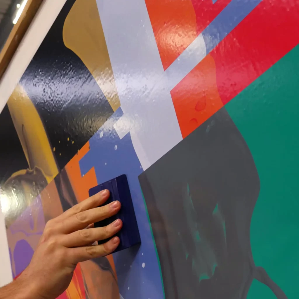 M12-P-P gloss white self-adhesive print vinyl being applied with a felt-edged squeegee onto a flat surface. The printed design is of an abstract set of wave shapes and water splash imagery set within brightly coloured boxed shapes that merge into each other.