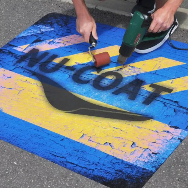 M15-M MaxGRAB print vinyl for self-adhesive graphic applications onto rough external surfaces such as onto tarmac, concrete, bricks, asphalt etc is pictured here in the process of being heat applied and rollered onto a tarmaced surface for the purpose of promotional signage with this printed floor graphic featuring the NU-COAT logo on a blue and yellow abstract cracked style pattern.
