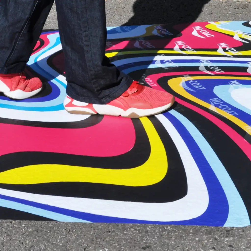 M15-M MaxGRAB self-adhesive floor print vinyl printed with a bright coloured swirling patterned promotional print that has been applied to an outdoor tarmac surface and is being walked over by a person wearing bright red trainers.