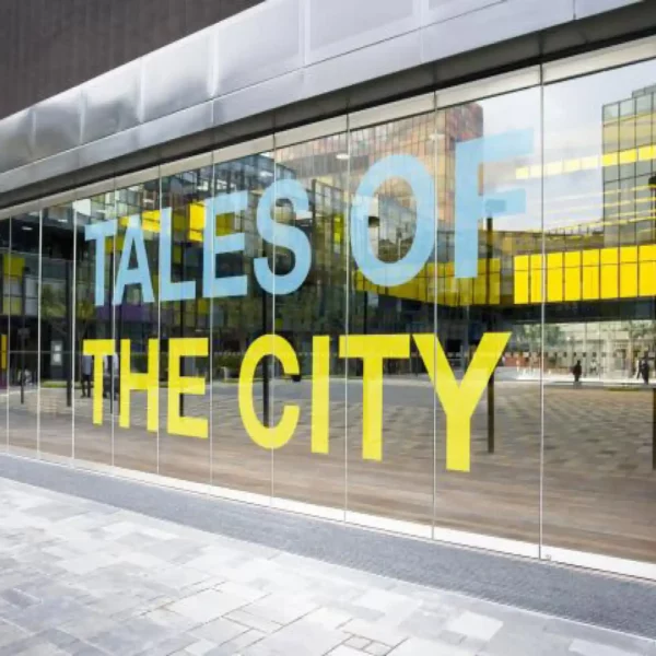 MACal 8300 cut letter window signage in pale blue and pale yellow. The vinyl sign spells out "Tales of the City" and the sign has been applied to a large external window that is reflecting the wider city in front of it.