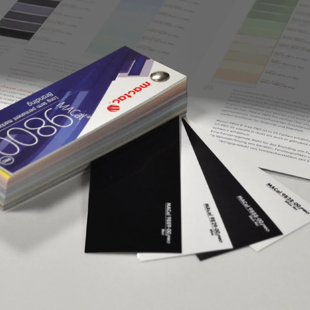 In the foreground of this photo, Mactac's MACal 9800 swatch is folded into a block with only the High Tack (9800 HT) black and white, gloss and matt vinyl samples sticking out. In the background is an opened colour card of the full range of 9800 coloured self-adhesive sign films, where just a few of the other colours that are also available in this swatch are faded into the back of the photo.