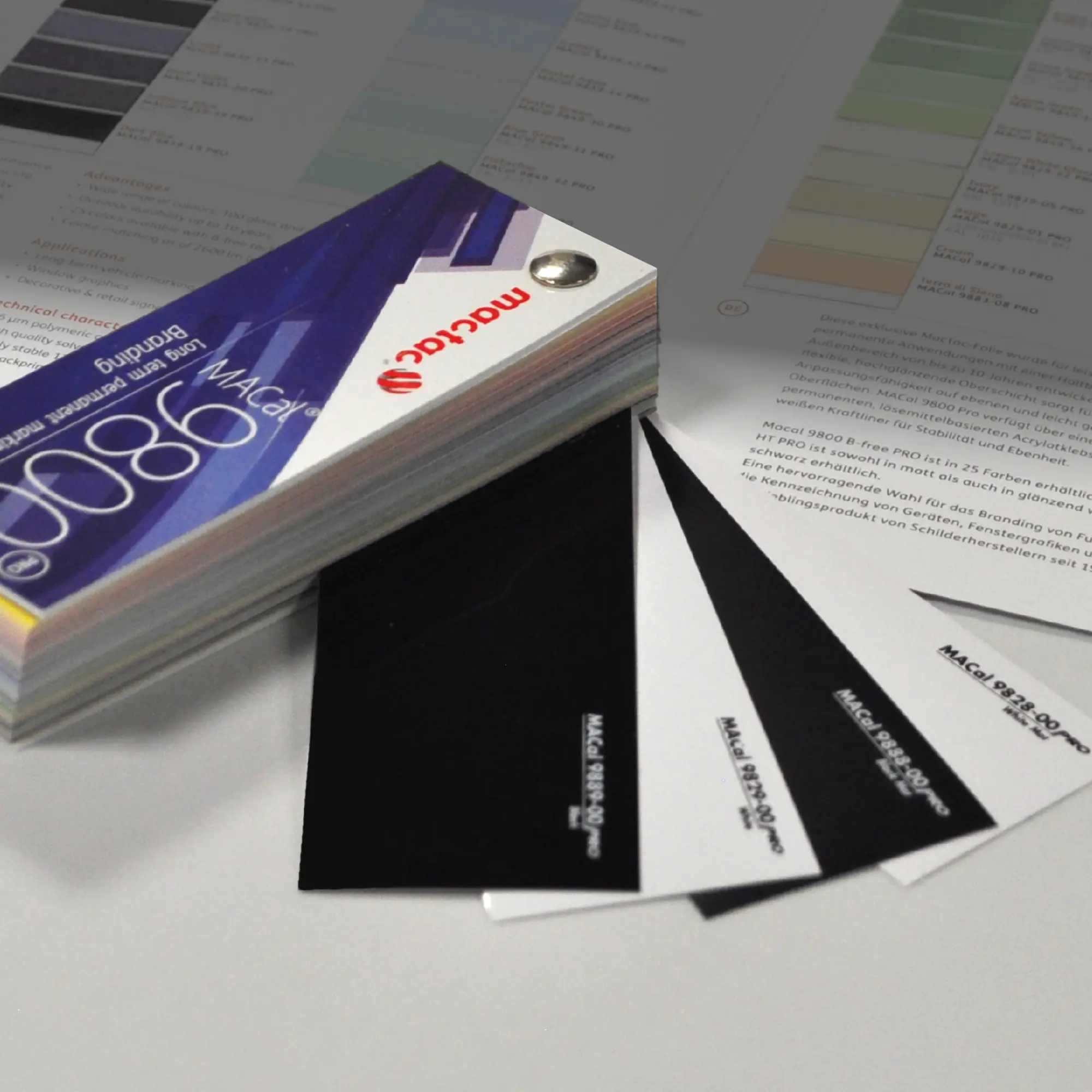 In the foreground of this photo, Mactac's MACal 9800 swatch is folded into a block with only the High Tack (9800 HT) black and white, gloss and matt vinyl samples sticking out. In the background is an opened colour card of the full range of 9800 coloured self-adhesive sign films, where just a few of the other colours that are also available in this swatch are faded into the back of the photo.