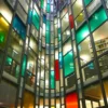 Mactac MACcrystal 8400 Series coloured window films in an abstract pattern of green, red and blue transparent tints applied to the glass windows on all floors in this office atrium, viewed from ground level and looing up, the dark grey edges of the glazing frame the coloured windows and make a striking, elegant and eye-catching colourful display.