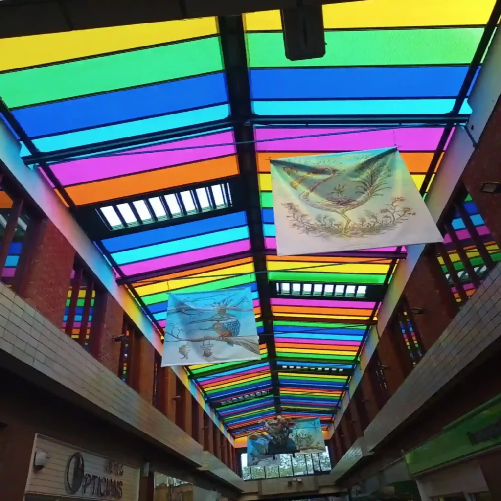 Mactac MACcrystal 8400 Series decorative transparent coloured window films applied to all of the glass panelled roof skylights in a shipping centre using almost the full spectrum of colours available in the range, allowing sunlight into the public area but with a tinted transparent effect which transforms the bright coloured ambience of the space.