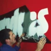 With the paint dried the paint mask stencil film is being peeled away as waste, leaving behind only the clean, crisp painted image on the red curtain side of this truck.