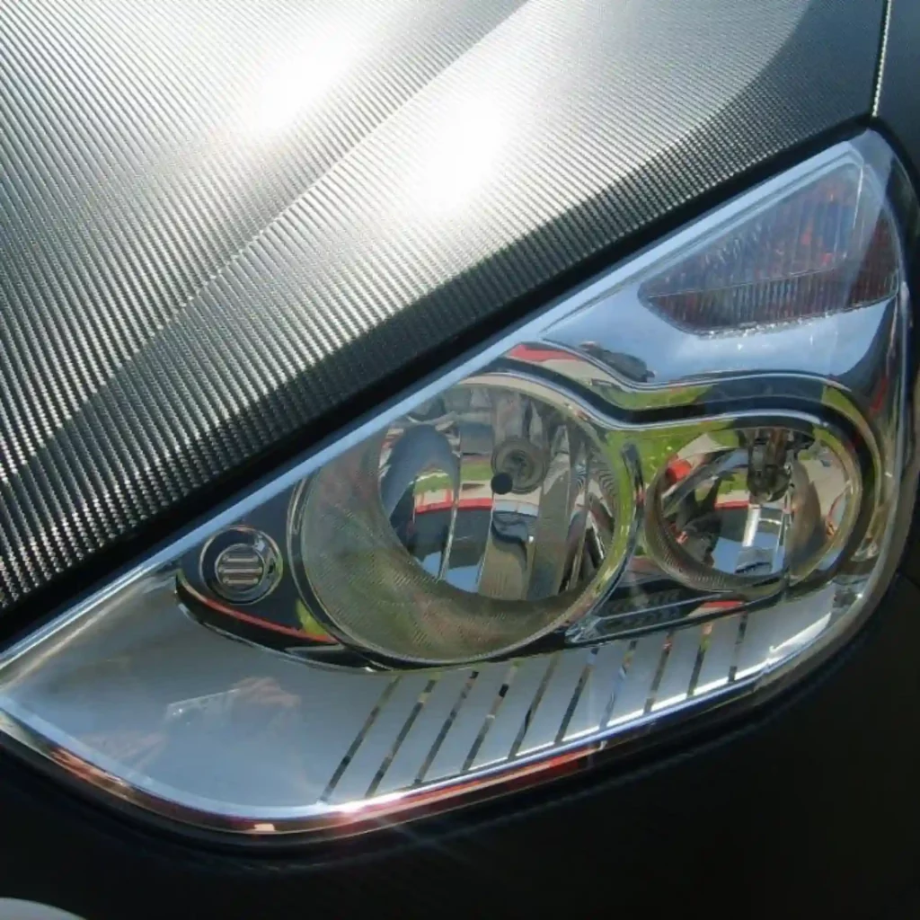 A close-up look at the detail of the carbon fibre pattern of Mactac's Black Carbon BF Tuning Film wrappping vinyl which has been applied to decorate the bonnet and around the headlight area of a modern road car, showing off the perfectly straight application of the embossed film with no warping of the carbon-fibre effect.